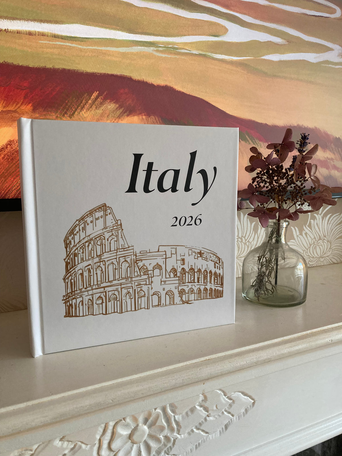 A hardcover There & Back Editions travel photo book standing on a white mantelpiece next to a small glass vase with flowers. A warm-toned abstract landscape painting is visible in the background.