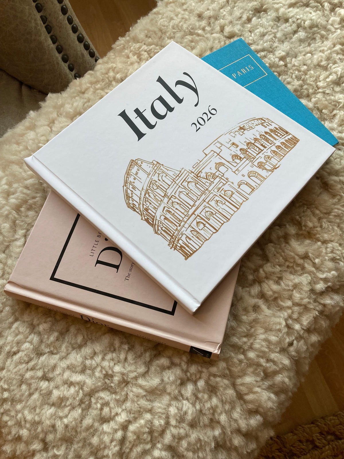 A stack of hardcover books and journals resting on a textured cream-colored rug. On the top is a travel photo book from There & Back Editions