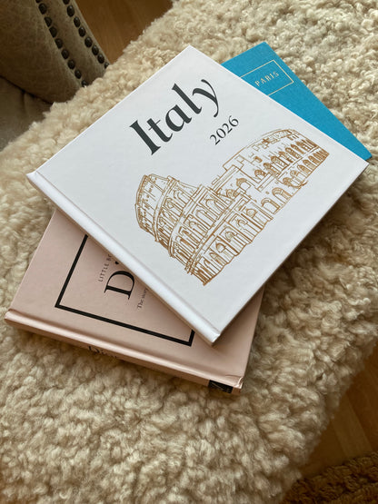 A stack of hardcover books and journals resting on a textured cream-colored rug. On the top is a travel photo book from There & Back Editions